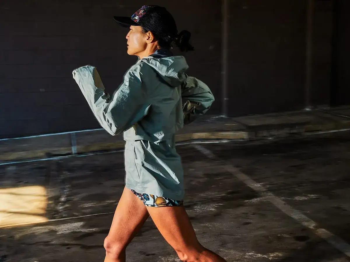 Donna corre sotto la pioggia, con giacca leggera e cappellino. L'asfalto bagnato riflette la luce.