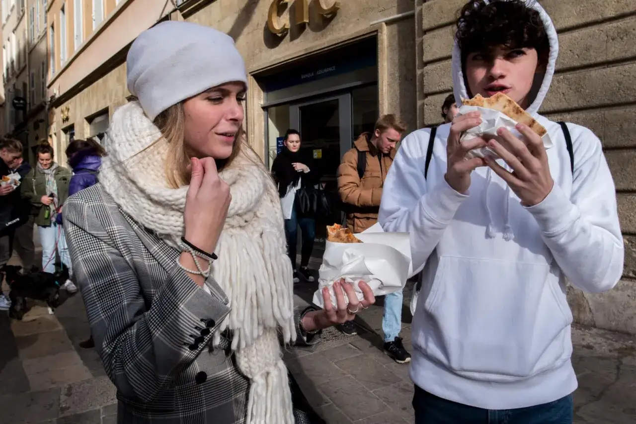 En ung kvinna med vit m&ouml;ssa och stor halsduk tittar p&aring; en kille som &auml;ter pizza. En scen f&aring;ngad med en Ricoh GR IV.