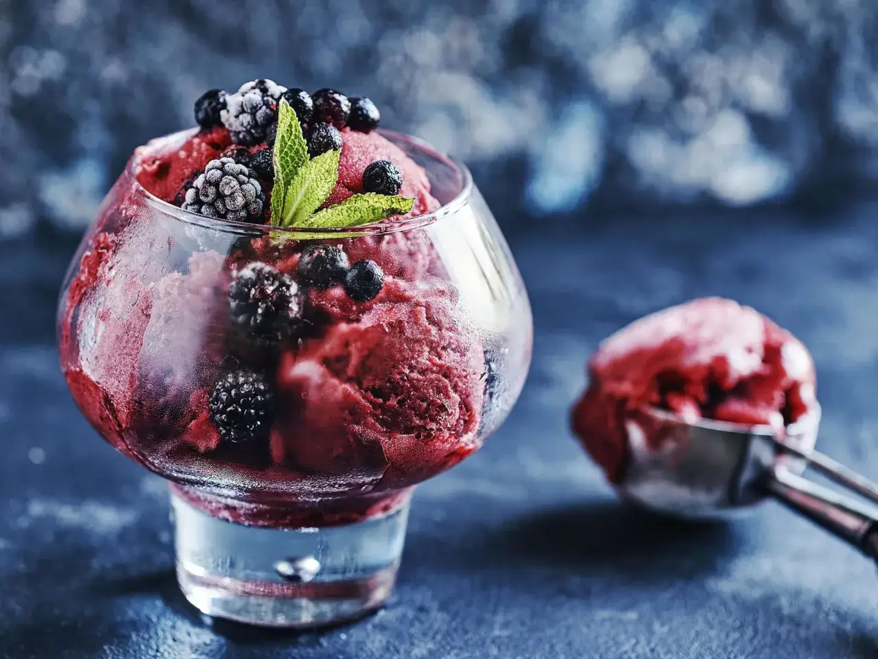 Ein Glas mit selbstgemachtem Brombeereis, garniert mit frischen Brombeeren und Minze. Daneben eine Eiskugel im Löffel.