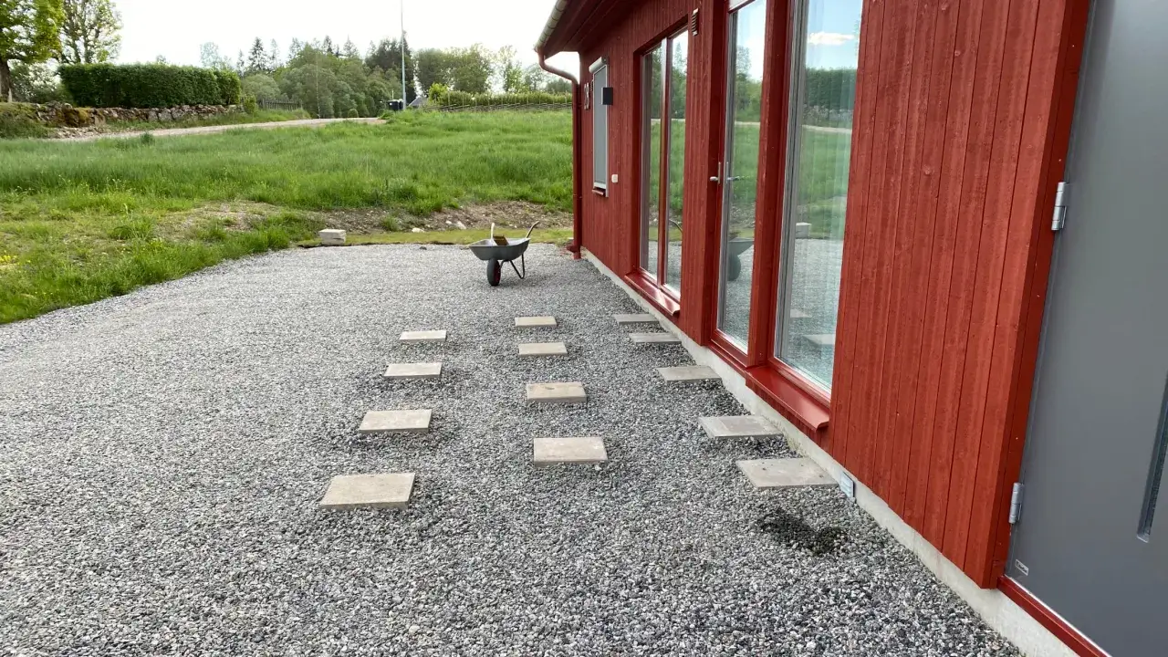 Stigande plattor leder från ett rött hus över grus. En skottkärra står redo för att hjälpa till med altanbygget.