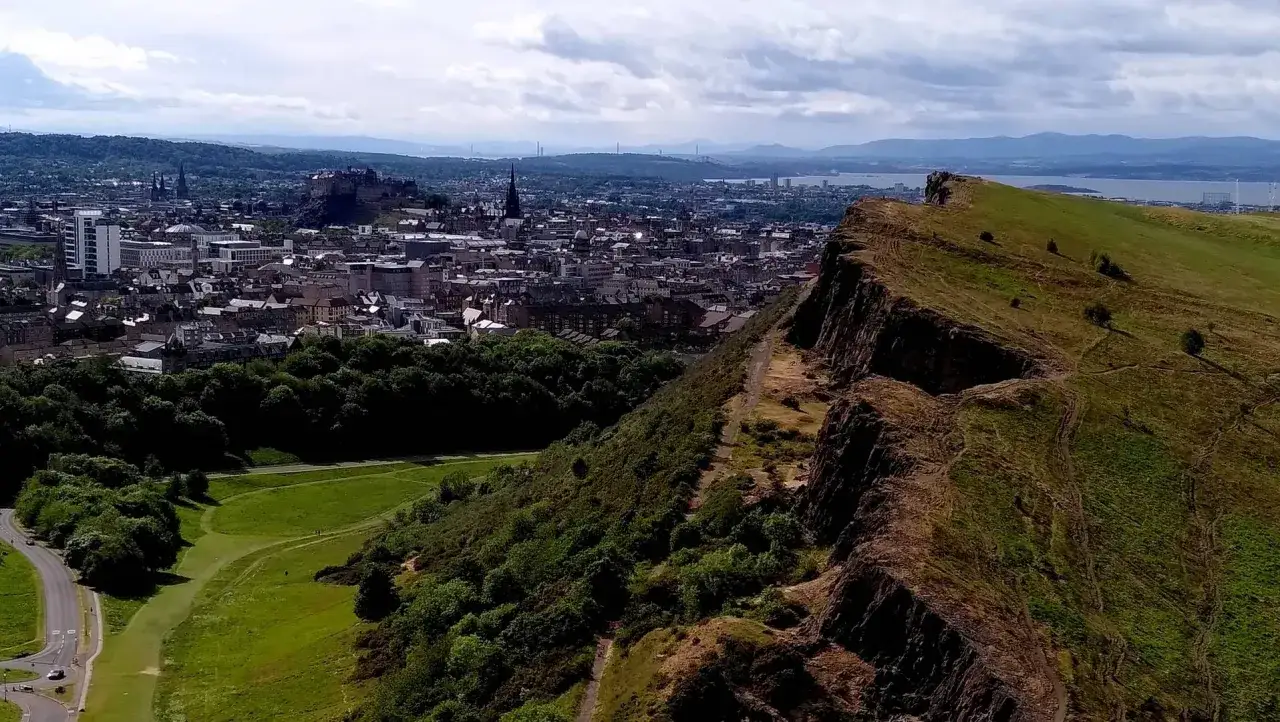 Vy över Skottlands huvudstad Edinburgh med dess slott på en kulle. En grönskande sluttning leder ner mot staden.
