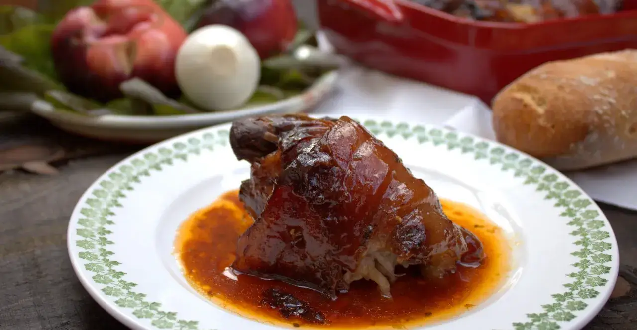 Codillo de cerdo glaseado, jugoso y dorado, servido en un plato blanco con borde verde. Acompa&ntilde;ado de pan y frutas.