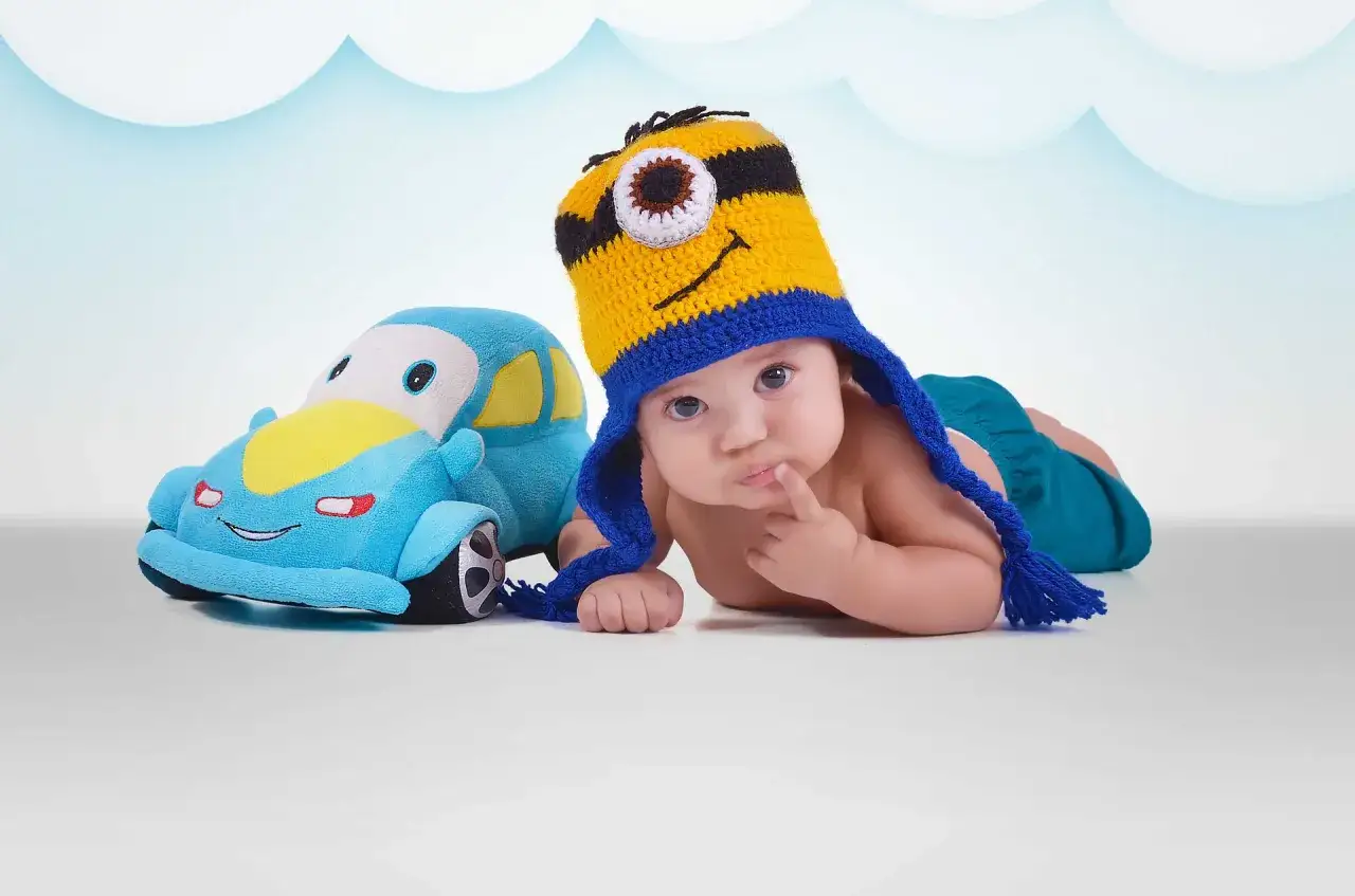 Maluch w czapce Minionka obok pluszowego autka, zastanawia się, kiedy dziecko zaczyna gaworzyć.