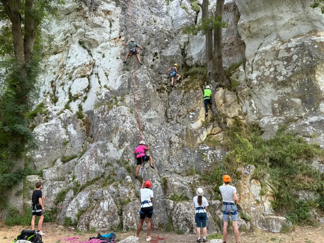 Des jeunes en pleine colonie de vacances escalade s'entraînent sur un mur rocheux, encordés et casqués, sous le regard de leurs accompagnateurs.