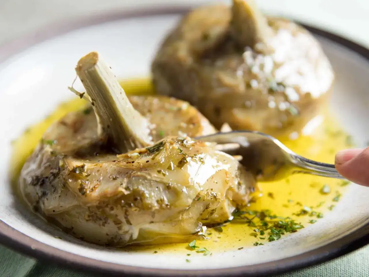Gegarte Artischocken in Öl mit Kräutern, ein köstliches Gericht für Rezepte mit frischen Artischocken. Eine Gabel sticht in eine Artischocke.
