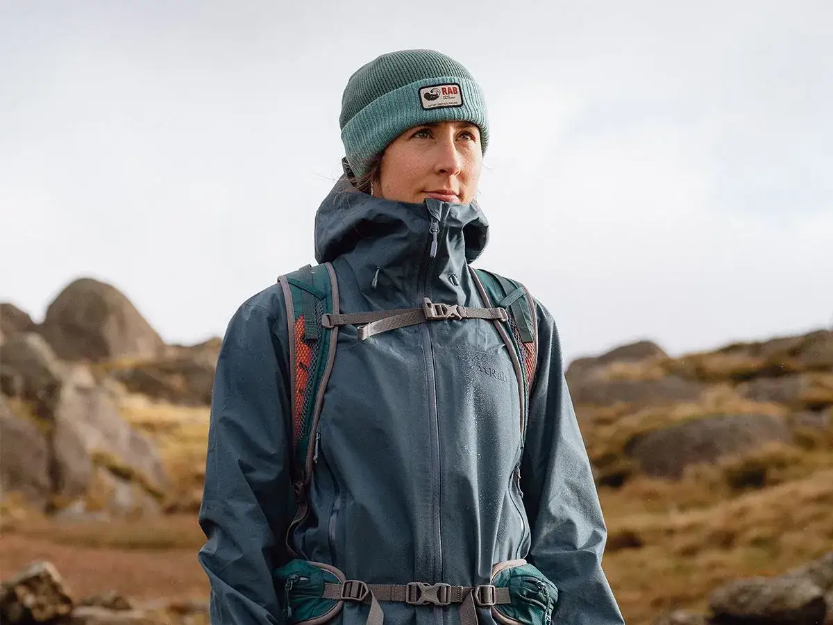 Frau in GORE-TEX INFINIUM Jacke und M&uuml;tze blickt nachdenklich in die Ferne, w&auml;hrend sie in felsiger Landschaft wandert.