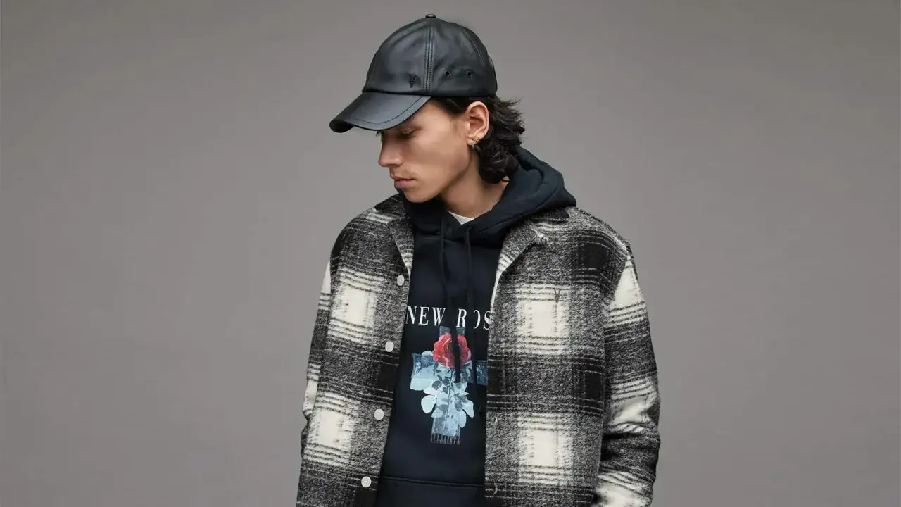 Mann mit schwarzer Lederkappe trägt ein kariertes Hemd über einem Hoodie mit Rosenmotiv. Perfekt, um ein Hemd mit Hoodie zu kombinieren.