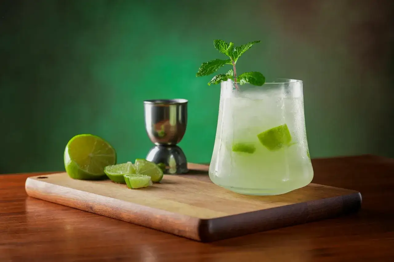 Orzeźwiający drink z limonką, lodem i miętą, obok pokrojonych kawałków limonki na desce.