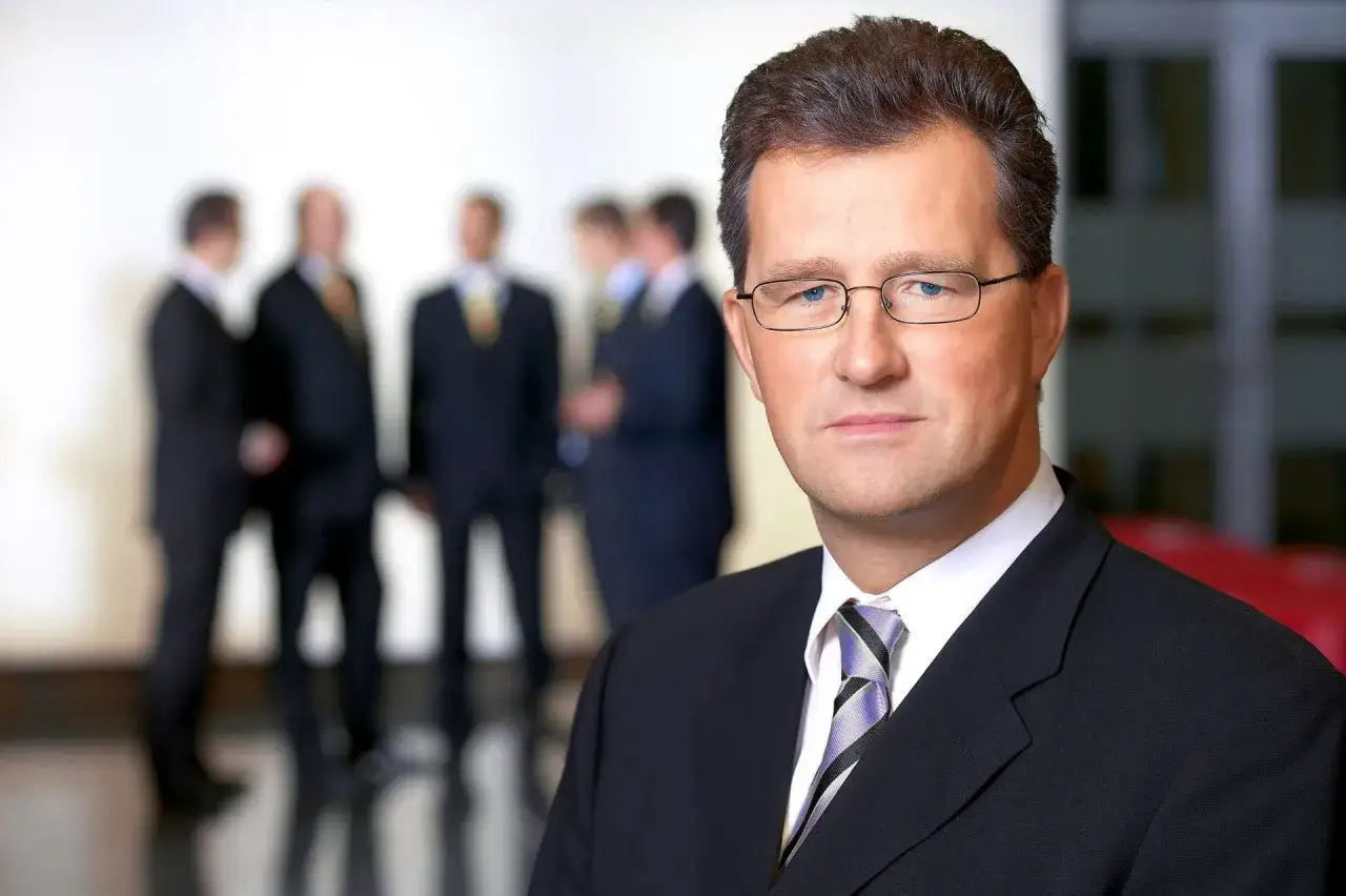 Ein Mann im Anzug mit Brille, im Hintergrund unscharf mehrere Männer. Er könnte ein Anwalt für Versicherung sein.