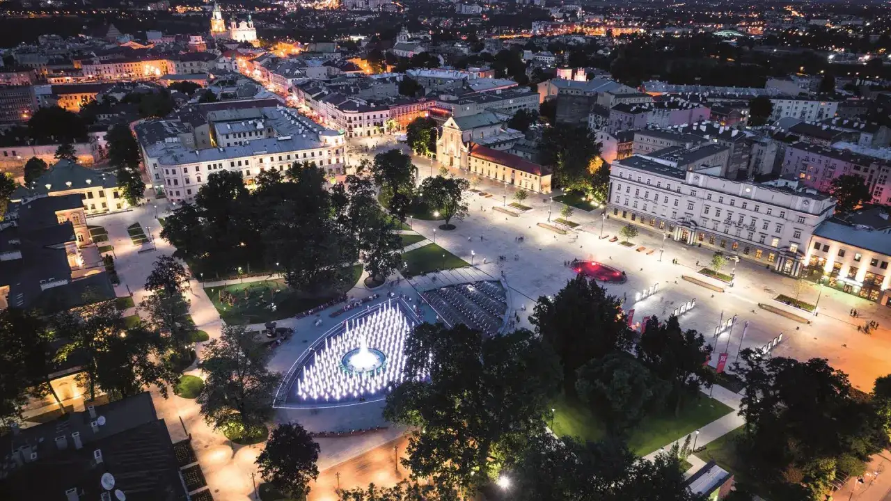 Nocny widok na plac Litewski z podświetlaną fontanną i otaczającą architekturą.