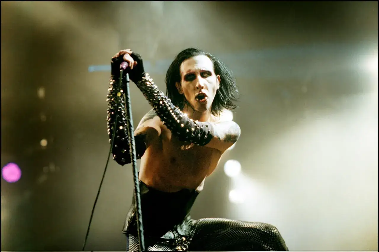 Marilyn Manson zespół na scenie, w charakterystycznym makijażu i kolczastych rękawiczkach, śpiewa do mikrofonu.