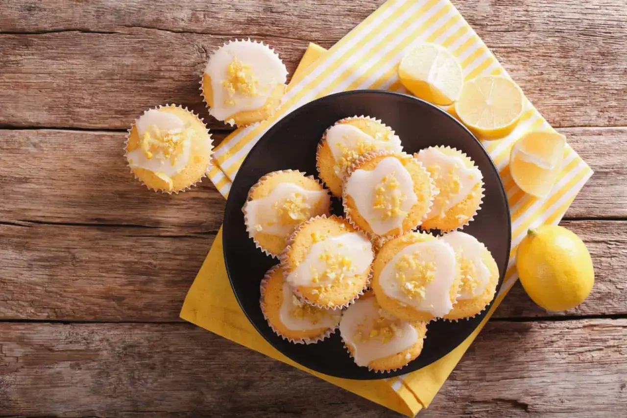 Zitronen-Muffins, frisch aus dem Thermomix, mit Zuckerguss und Zitronenschale bestreut, auf einem Teller mit Zitronenspalten.