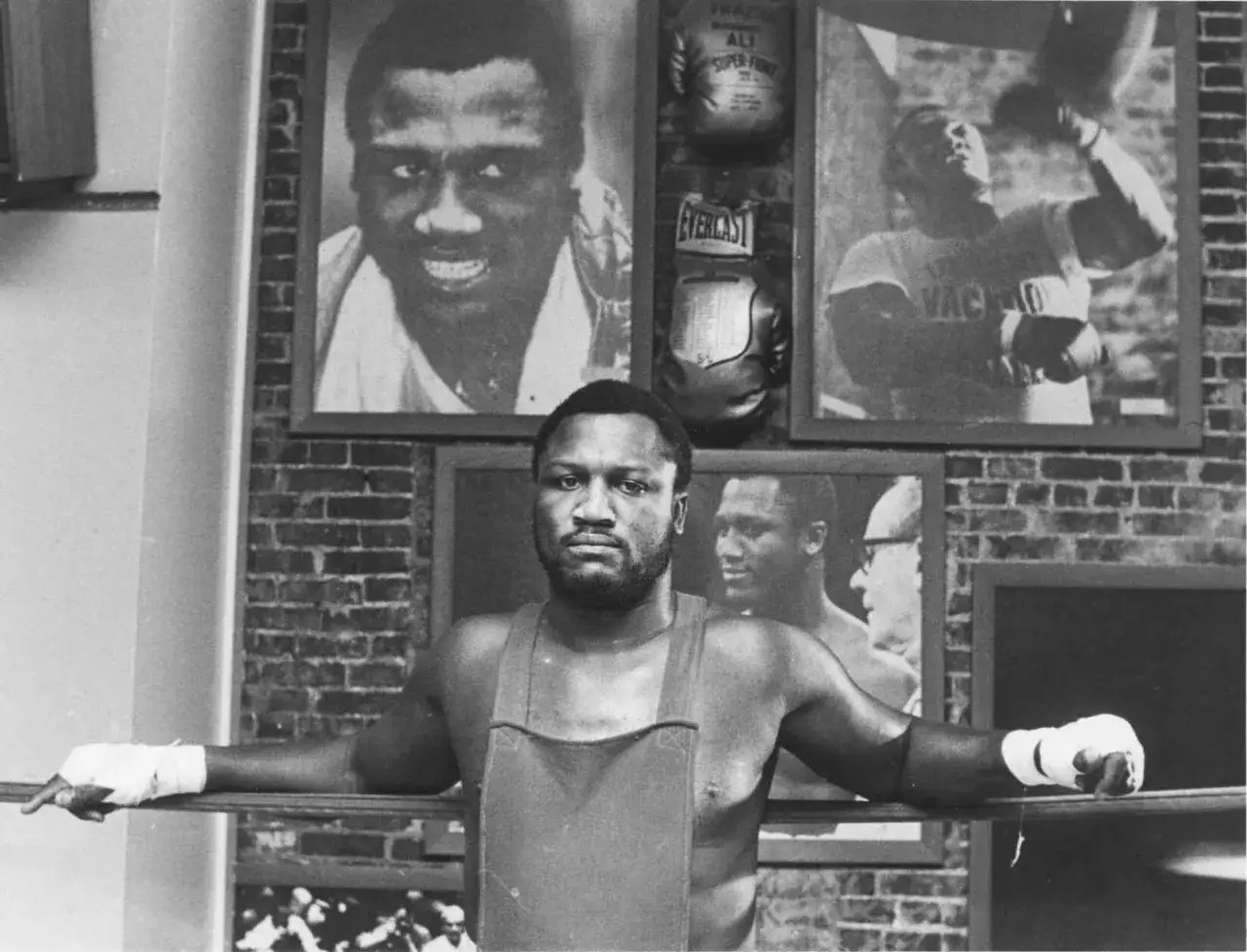 Joe Frazier, young and determined, leans on the ropes. Photos of his triumphs, including one with Ali, adorn the wall.