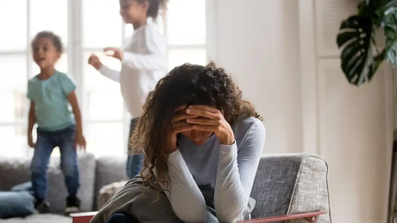 Frau mit Händen vor dem Gesicht, während Kinder im Hintergrund spielen. Ein Bild, das die Herausforderungen der **Pflege bei Depression** verdeutlicht.
