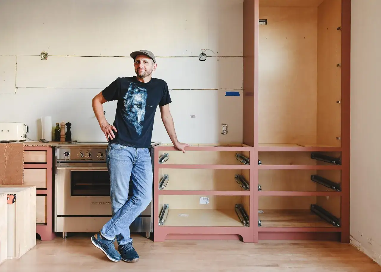 Man in wolf t-shirt stands by unfinished kitchen cabinets, ready to learn how to hang cabinets.