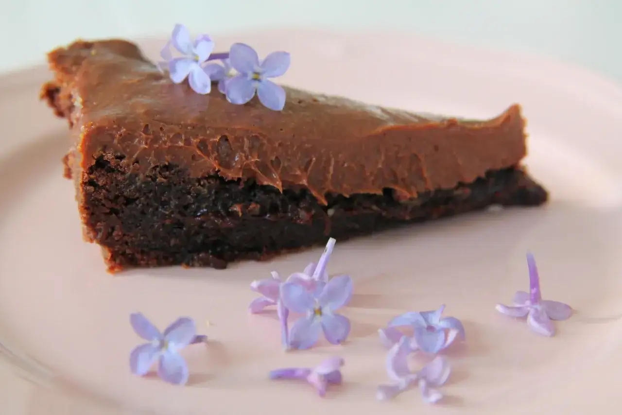 En bit chokladkaka med tryffel, dekorerad med små lila blommor på en ljusrosa tallrik.