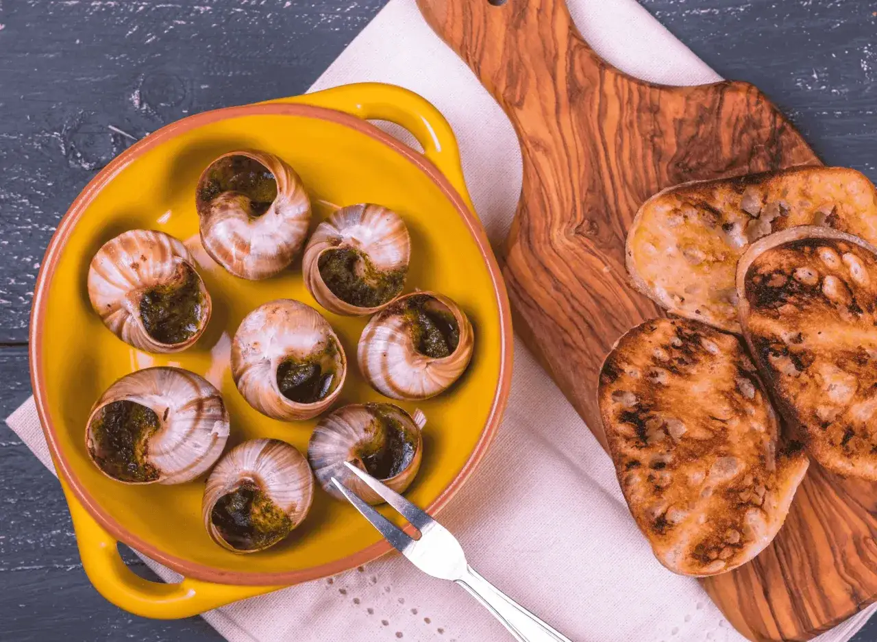 Co to jest escargot? Siedem ślimaków w muszlach z zielonym nadzieniem w żółtej misce, obok grzanki na desce z oliwnego drewna.