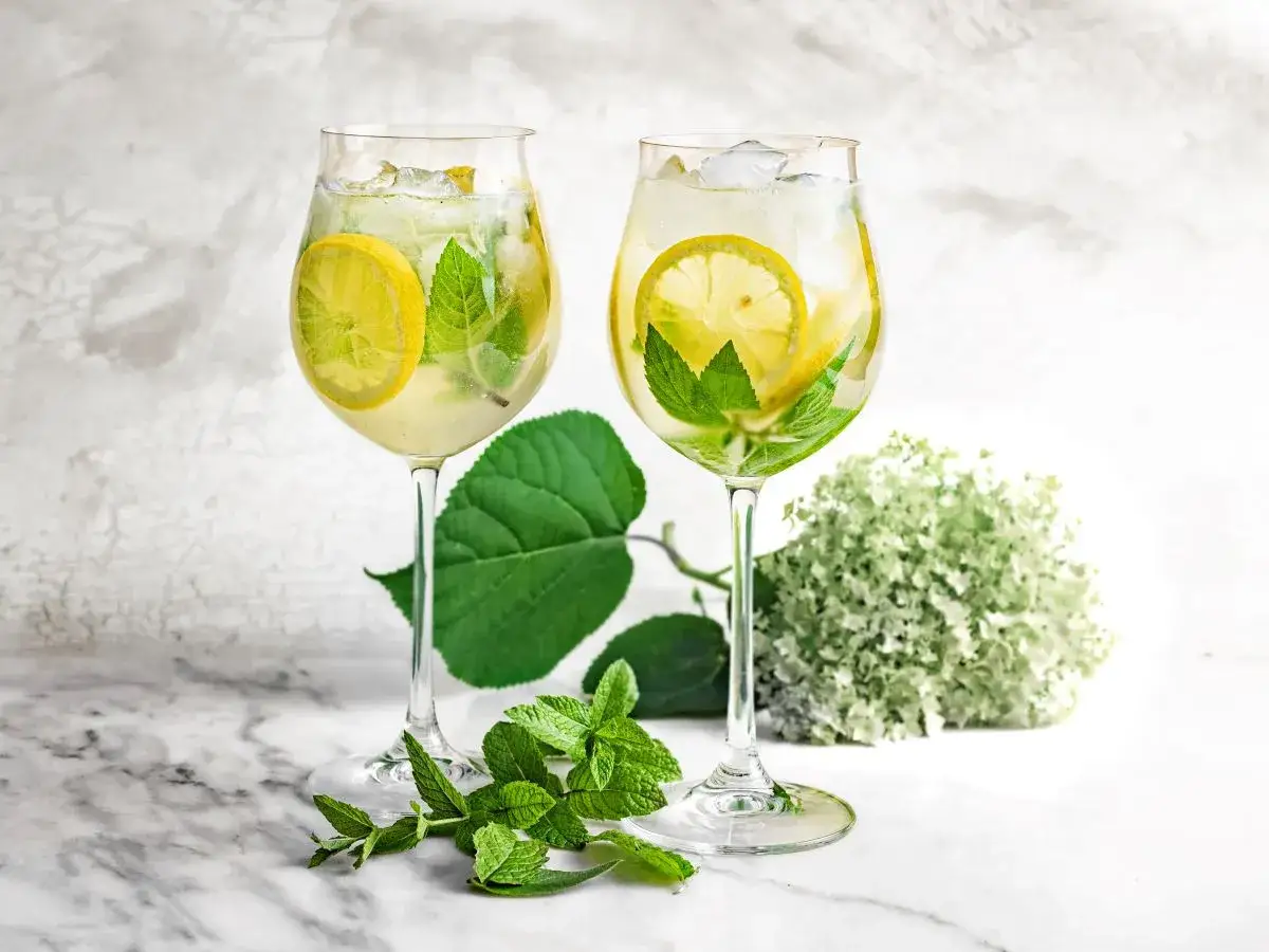 Orzeźwiający drink z prosecco i sokiem z bzu, ozdobiony plasterkami cytryny i listkami mięty, z kwiatami bzu w tle.