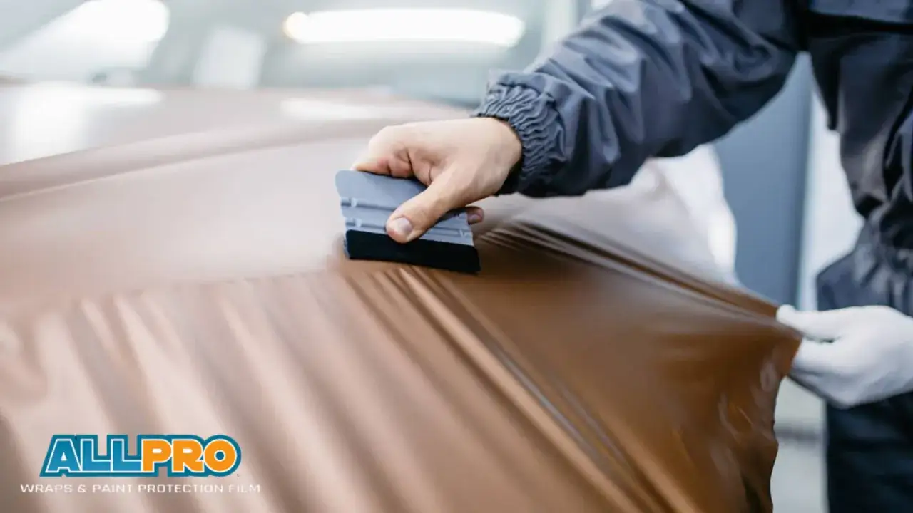 A person's hands, one with a squeegee, apply a metallic brown vinyl wrap to a car. This DIY car wrap project is in progress.