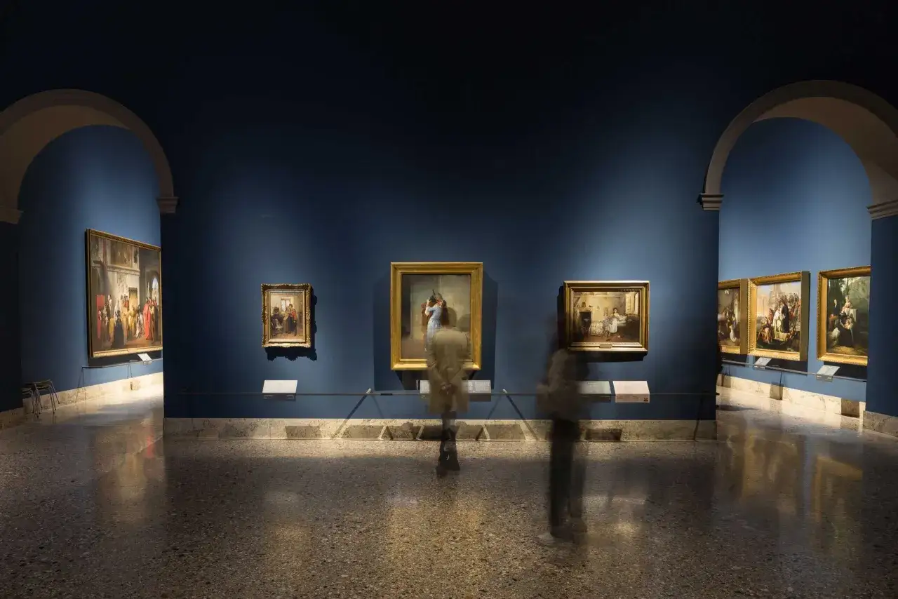 Wnętrze Pinacoteca di Brera z obrazami na niebieskich ścianach i rozmazanymi postaciami zwiedzających.