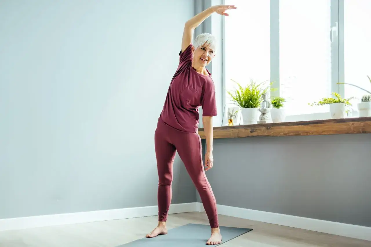 Une femme senior souriante pratique un exercice d'étirement latéral, un élément clé de son parcours équilibre senior, près d'une fenêtre lumineuse.