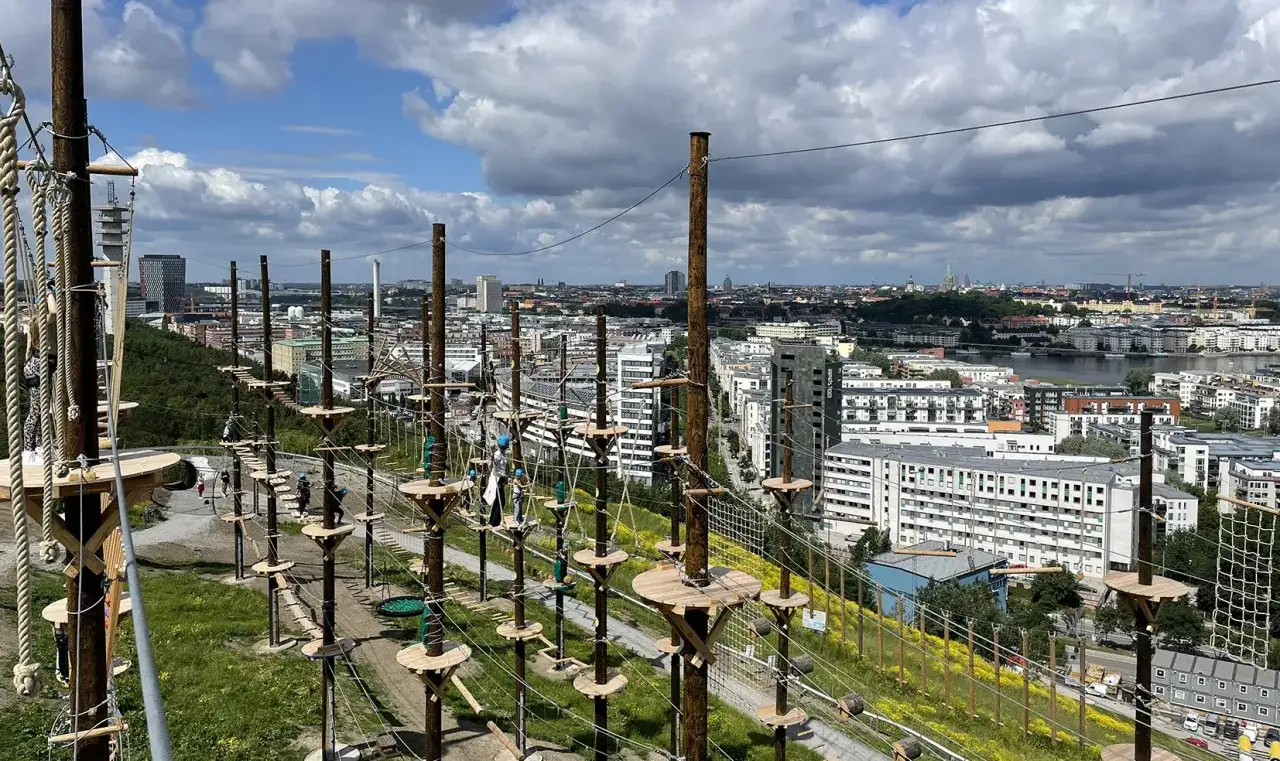 Hög höghöjdsbana vid Hammarbybacken med Stockholm i bakgrunden. Människor klättrar och balanserar på träplattformar och rep.