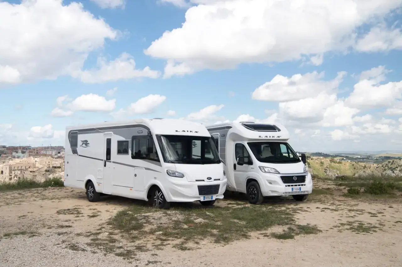 Deux camping-cars blancs de type intégral, marque Laika, stationnés sur un terrain rocailleux avec une ville en arrière-plan.