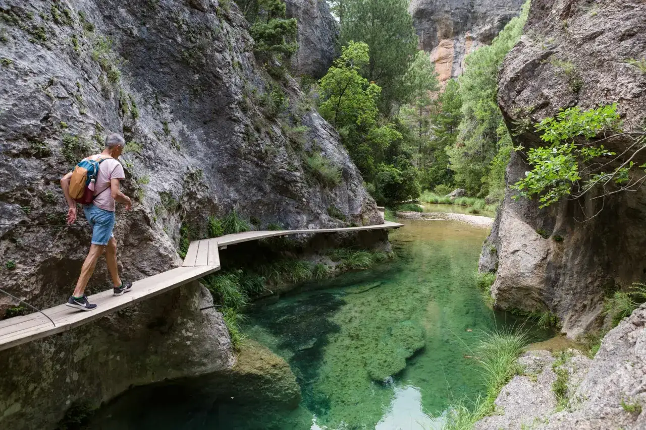 Hombre camina por pasarela de madera en la ruta parrizal de beceite, junto a un río de aguas cristalinas y paredes rocosas.