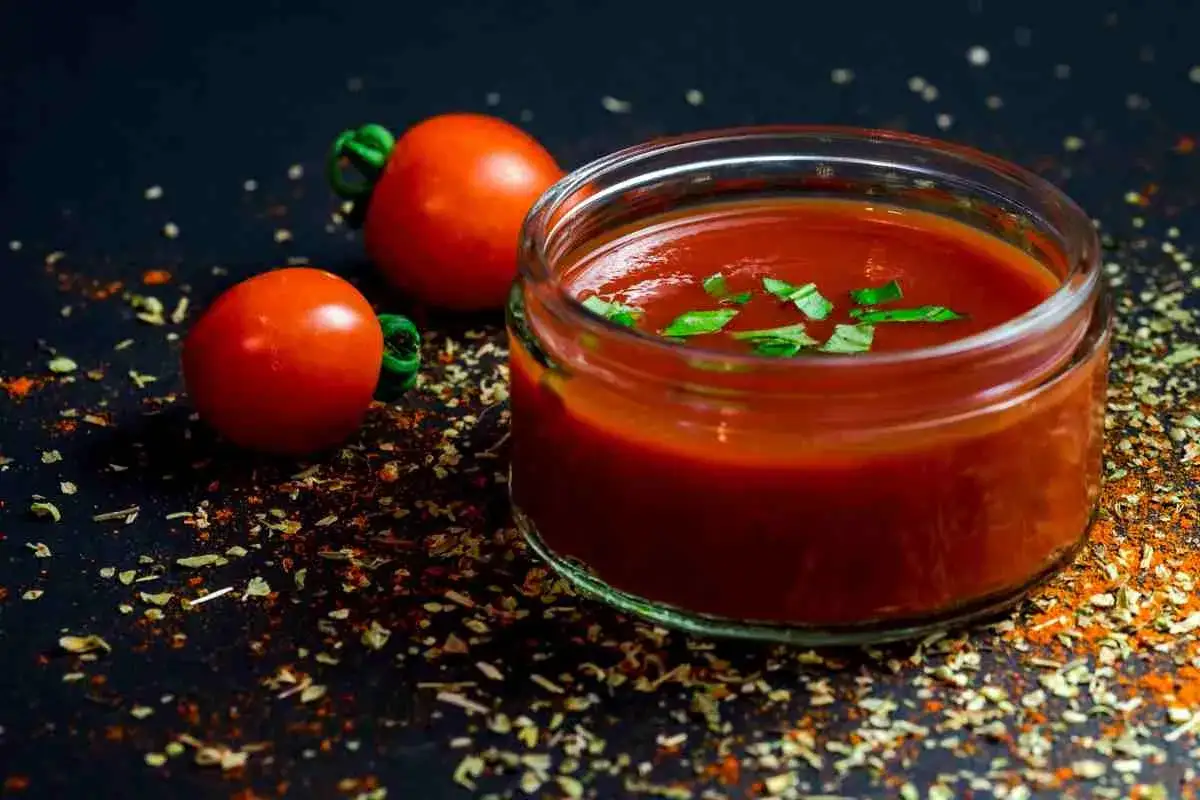 Un vasetto di ketchup fatto in casa, guarnito con basilico fresco, accanto a due pomodori maturi e spezie sparse.