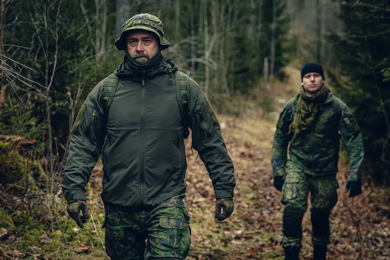 Dos hombres con equipo militar caminan por un bosque. Uno lleva una balaclava verde y un sombrero de camuflaje.