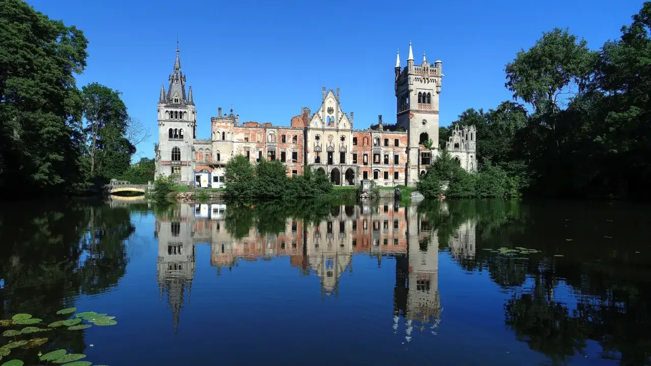 Zamek Kopice, ruiny zamku z wieżami, odbijające się w spokojnej tafli jeziora, otoczone zielenią.