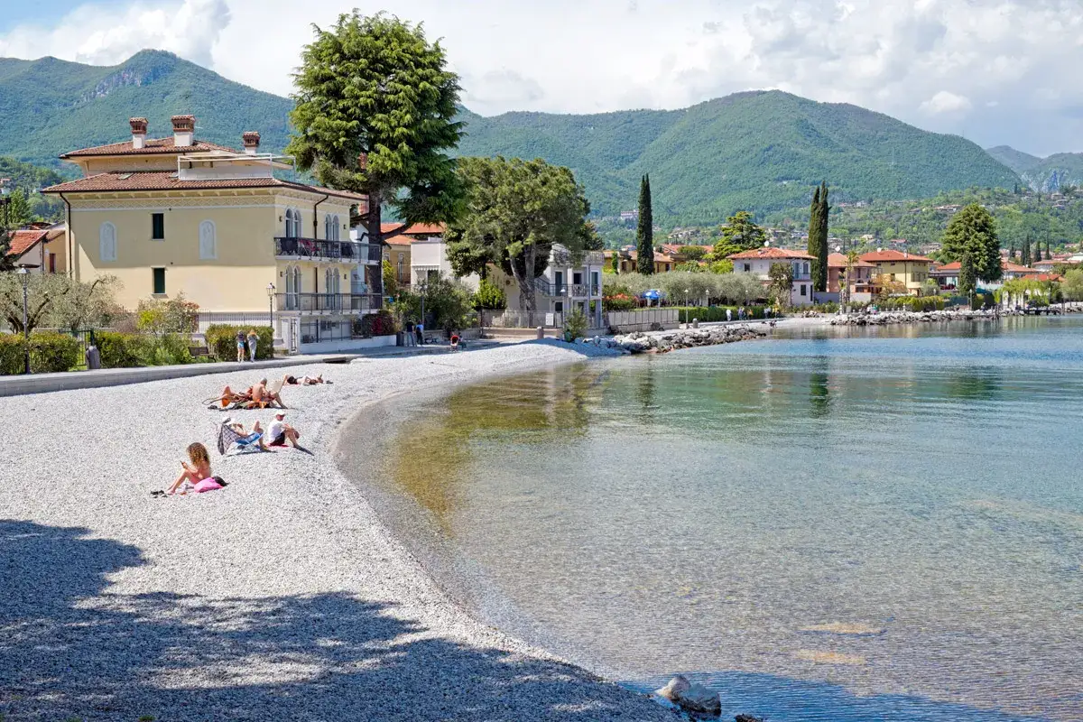 Spiaggia di ciottoli a San Michele Sal&ograve;, con persone che prendono il sole e case colorate sullo sfondo, sotto montagne verdi.
