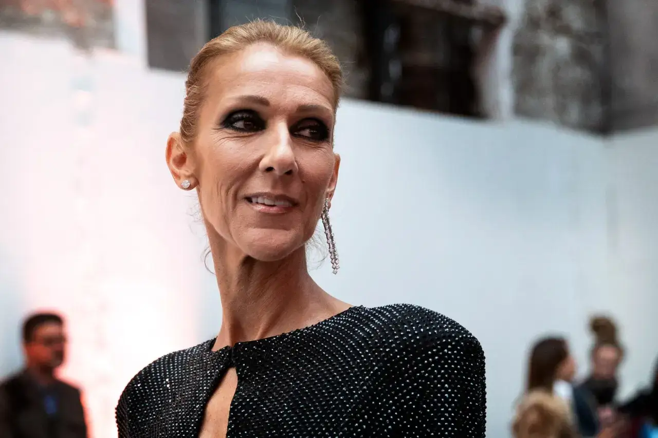 Celine Dion w błyszczącej sukni, z wyrazistym makijażem i długimi kolczykami. W tle widać zamazane sylwetki ludzi.