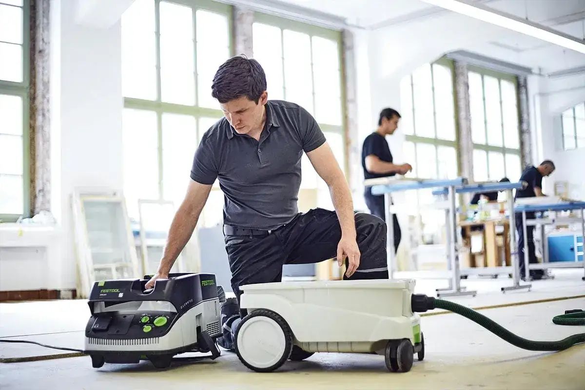 Un homme ajuste son aspirateur Festool CTL 36 E AC dans un atelier lumineux. D'autres travaillent en arri&egrave;re-plan.