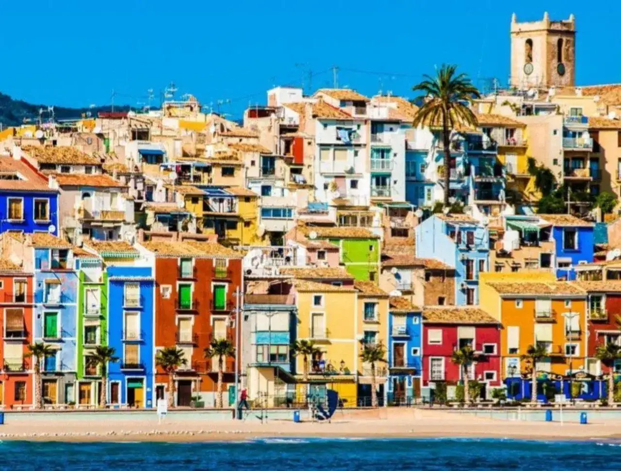Casas de colores vibrantes en la costa, un ejemplo de los pueblos bonitos de Alicante. Palmeras y el mar azul completan la escena.