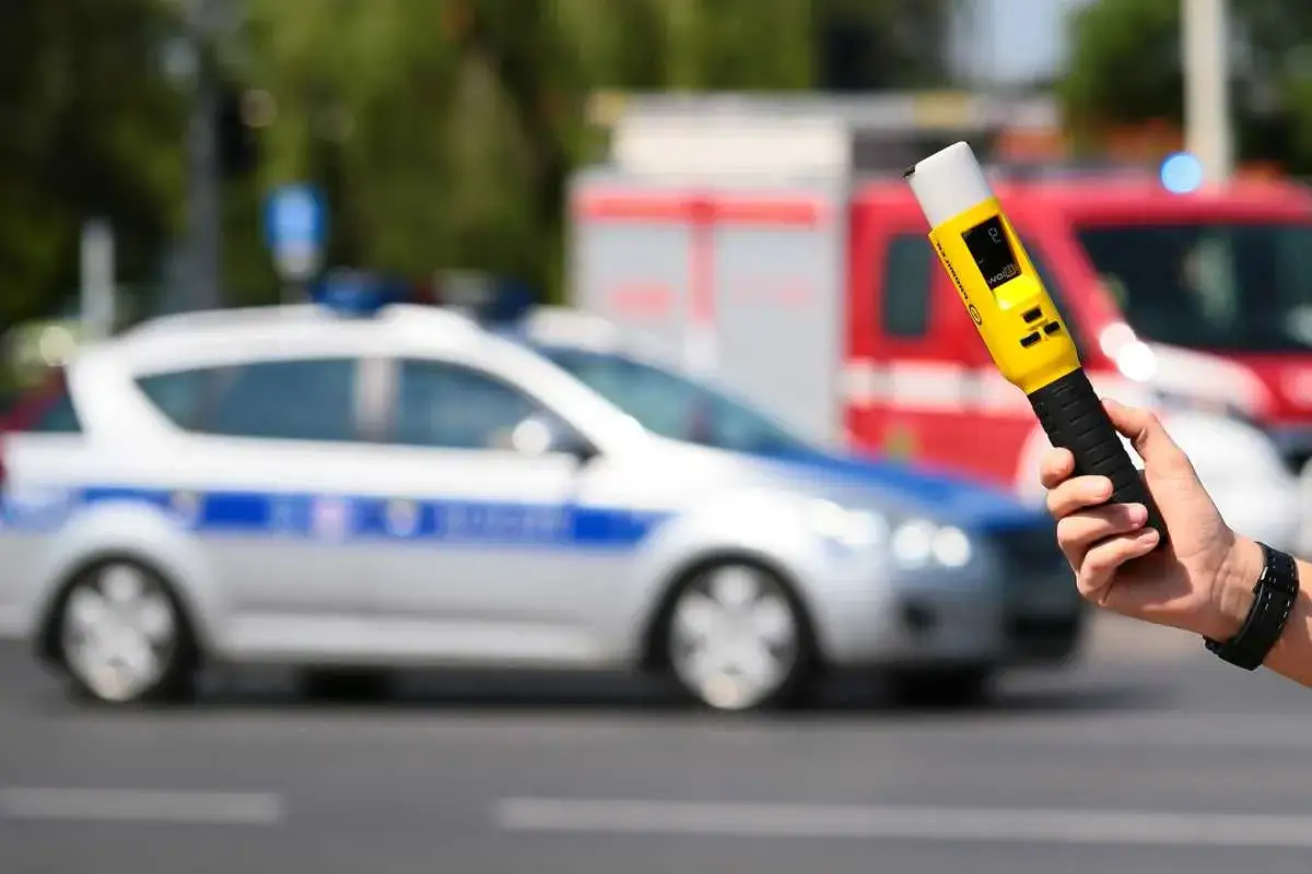 Ręka trzyma alkomat. W tle radiow&oacute;z i straż pożarna. Tylko kt&oacute;re badanie alkomatem się liczy, gdy bezpieczeństwo jest najważniejsze.