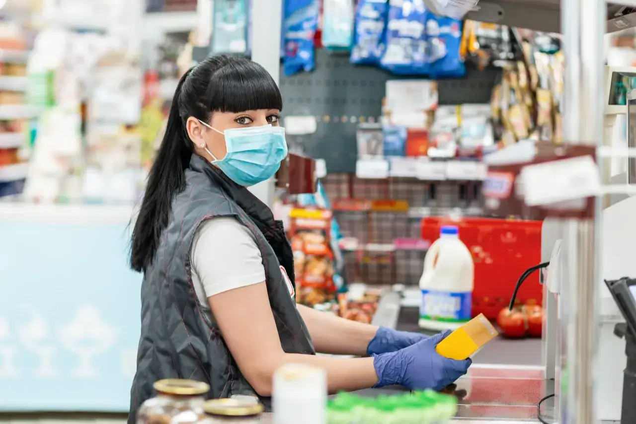 Cassiera con mascherina e guanti in un supermercato, mentre passa un prodotto al nastro. La GDO, grande distribuzione organizzata, è questo: un luogo di acquisto quotidiano.