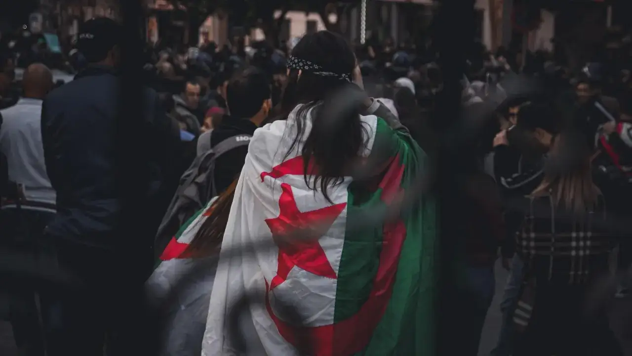 Eine Frau tr&auml;gt die algerische Flagge, ein Symbol der algerischen Kultur, &uuml;ber ihren Schultern. Sie ist Teil einer Menschenmenge, die sich auf einer Stra&szlig;e versammelt hat.