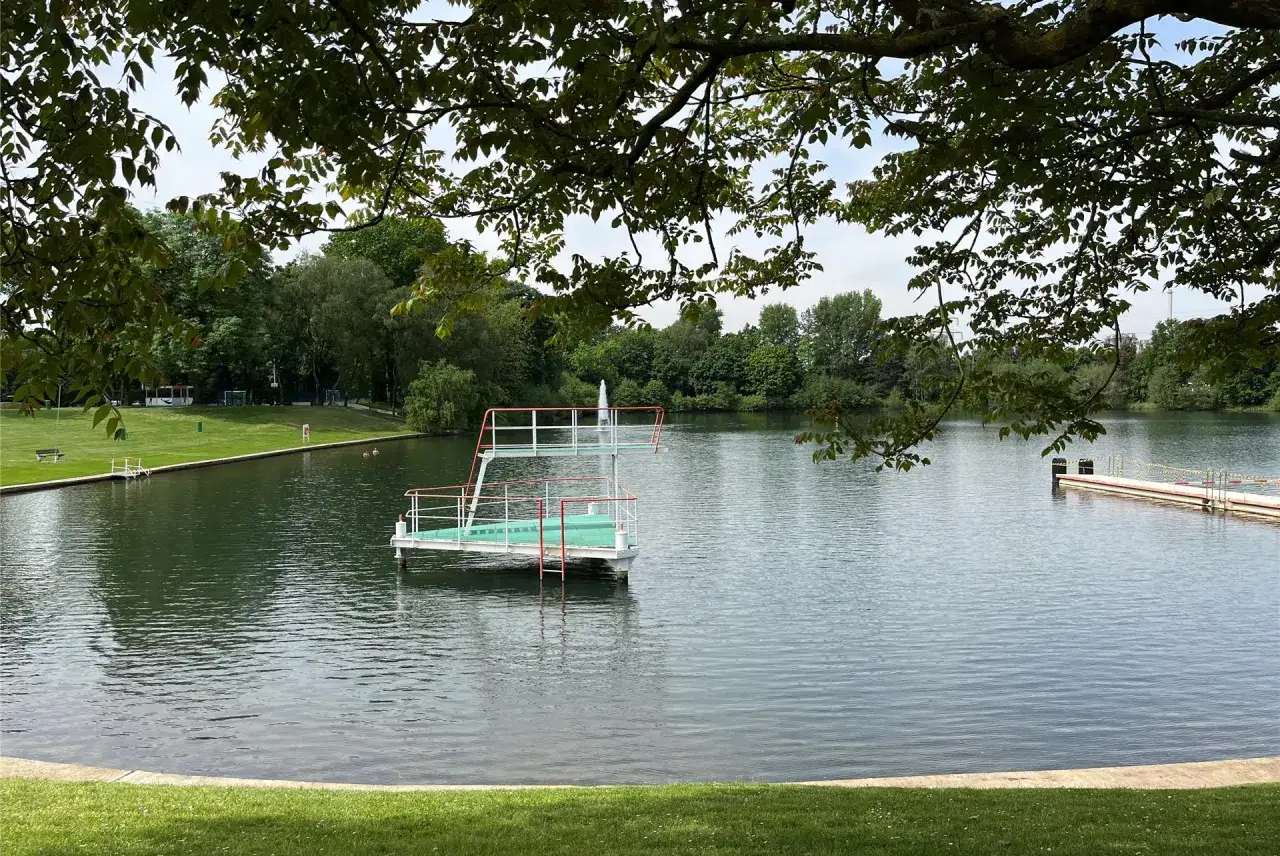Ein Sprungturm ragt aus dem Wasser des Badeweiher Marl. Im Hintergrund gr&uuml;ne Ufer und B&auml;ume.