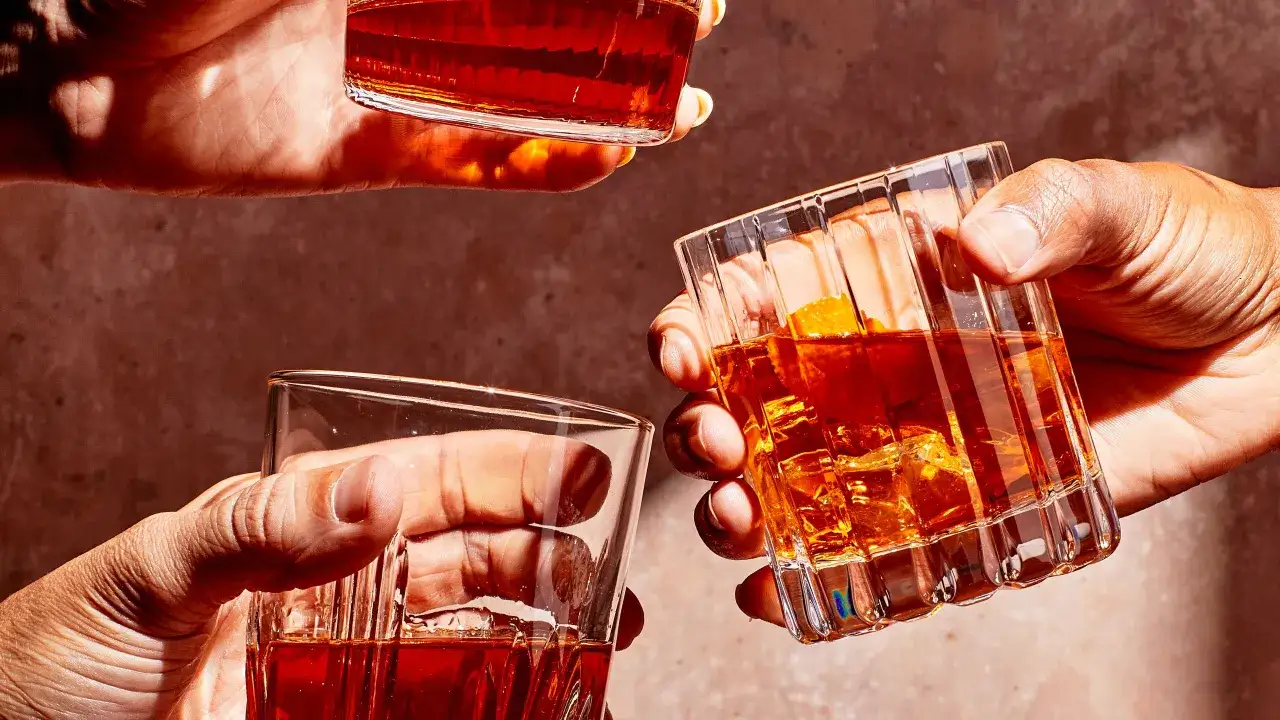 Drei Hände halten Gläser mit bernsteinfarbenem Whisky. Ein Glas enthält Eis und eine Orangenzeste, was auf einen Cocktail hindeutet, der den Unterschied zwischen Single Malt und Blended Whisky veranschaulicht.