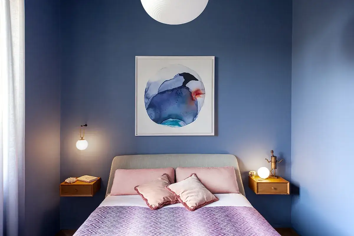 Camera da letto con pareti blu intenso, un quadro astratto e tessuti rosa cipria. I colori camera da letto cromoterapia creano un'atmosfera rilassante.