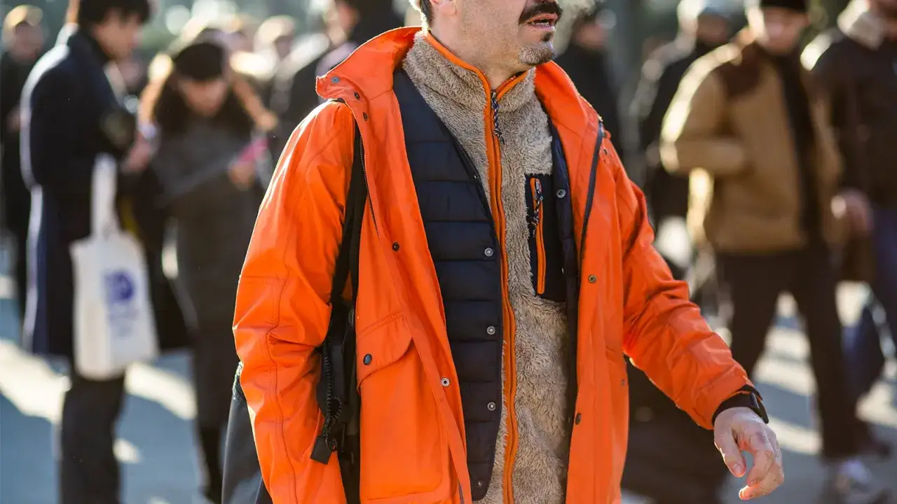 Mann im **gorpcore style** mit leuchtend oranger Jacke, schwarzer Weste und flauschigem Fleece.