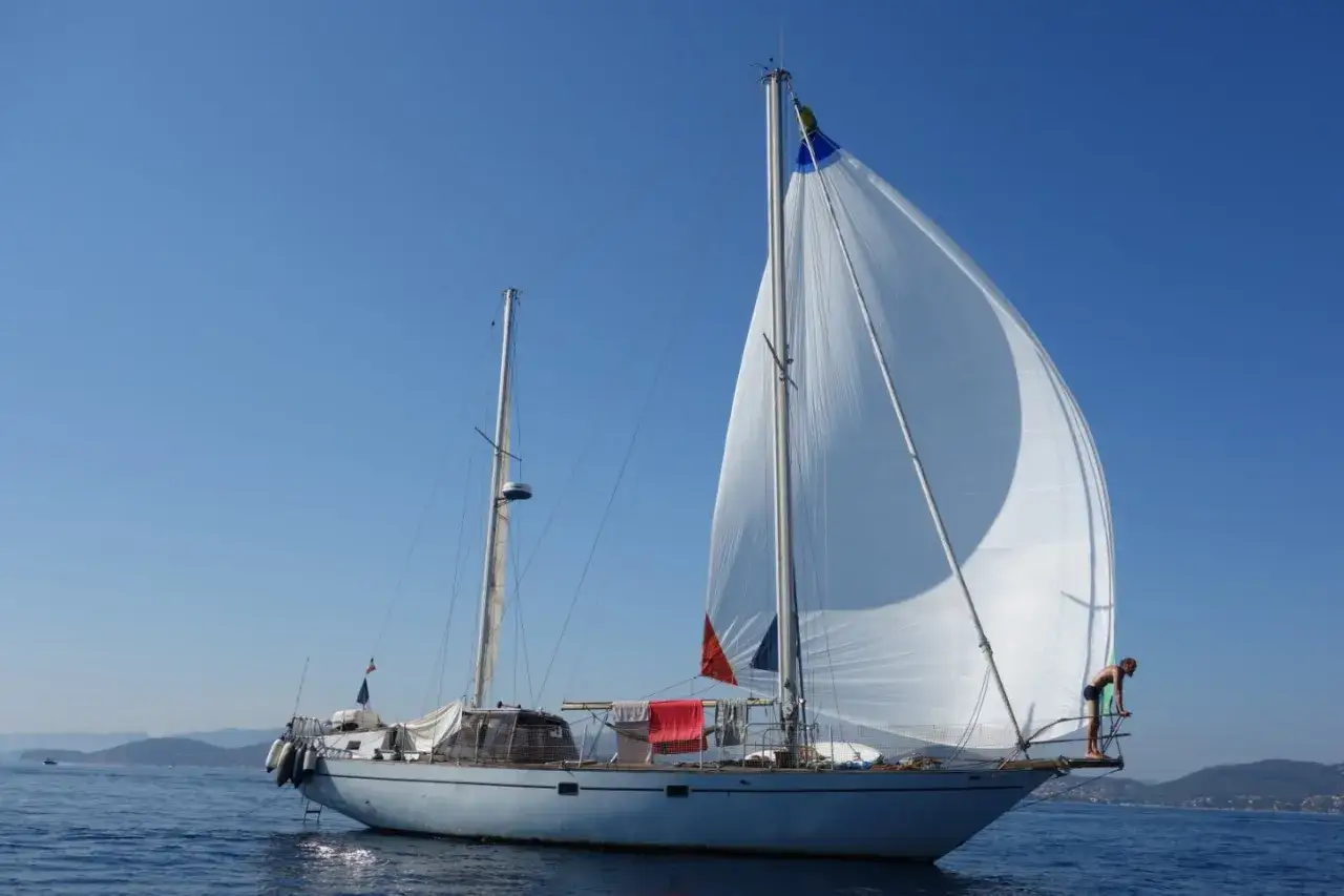 Un bateau corsaire blanc navigue sous un ciel bleu éclatant, ses voiles gonflées par le vent. Un homme est debout à la proue.