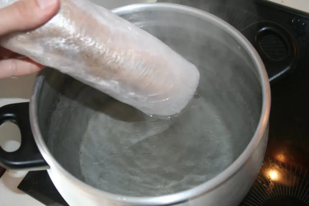 Une main plonge un rouleau de foie gras emball&eacute; dans de l'eau bouillante dans une casserole.