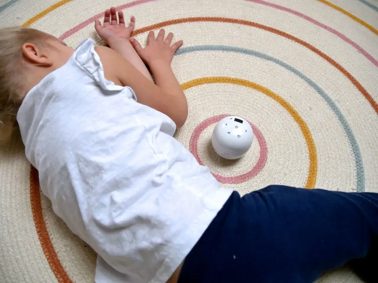 Kleines Kind liegt auf einem Teppich mit bunten Kreisen. Ein weißer Ball mit Knöpfen liegt daneben. Hilft reizoffene Kinder zur Ruhe zu kommen.