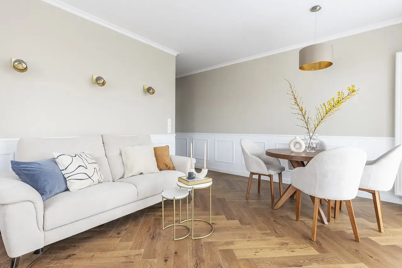 Przytulny salon z beżową kanapą, stolikami i okrągłym stołem. Na ścianie wiszą złote cokoliki, dodając elegancji.