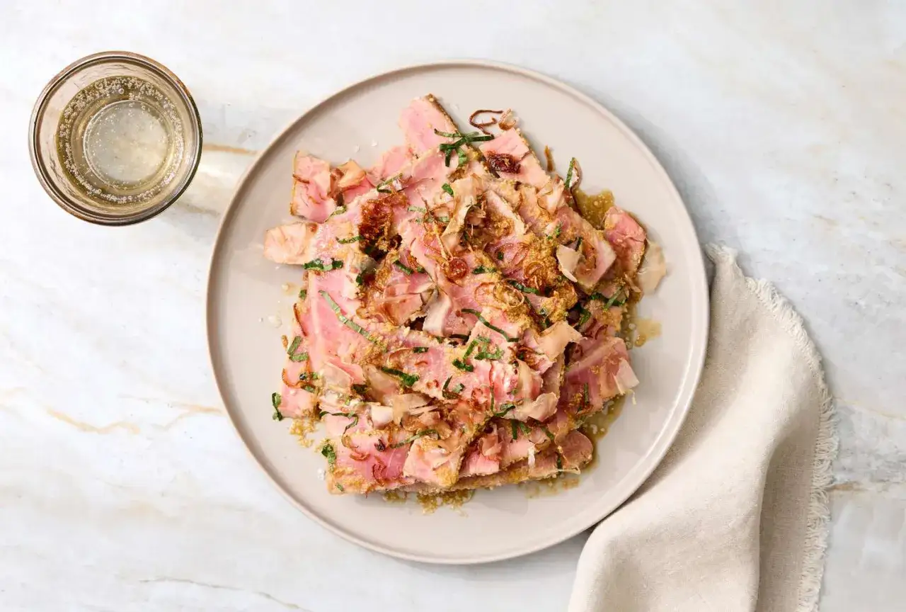 Tataki de at&uacute;n, finas l&aacute;minas de at&uacute;n sellado con un toque de hierbas y crujientes cebollitas fritas, servido en un plato con un vaso de bebida espumosa.