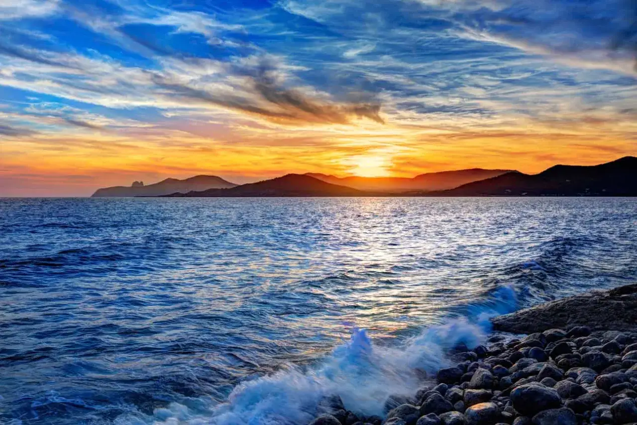 Coucher de soleil sur la mer, avec des vagues s'écrasant sur une plage de galets. Un paysage qui donne envie de savoir où aller à Ibiza.