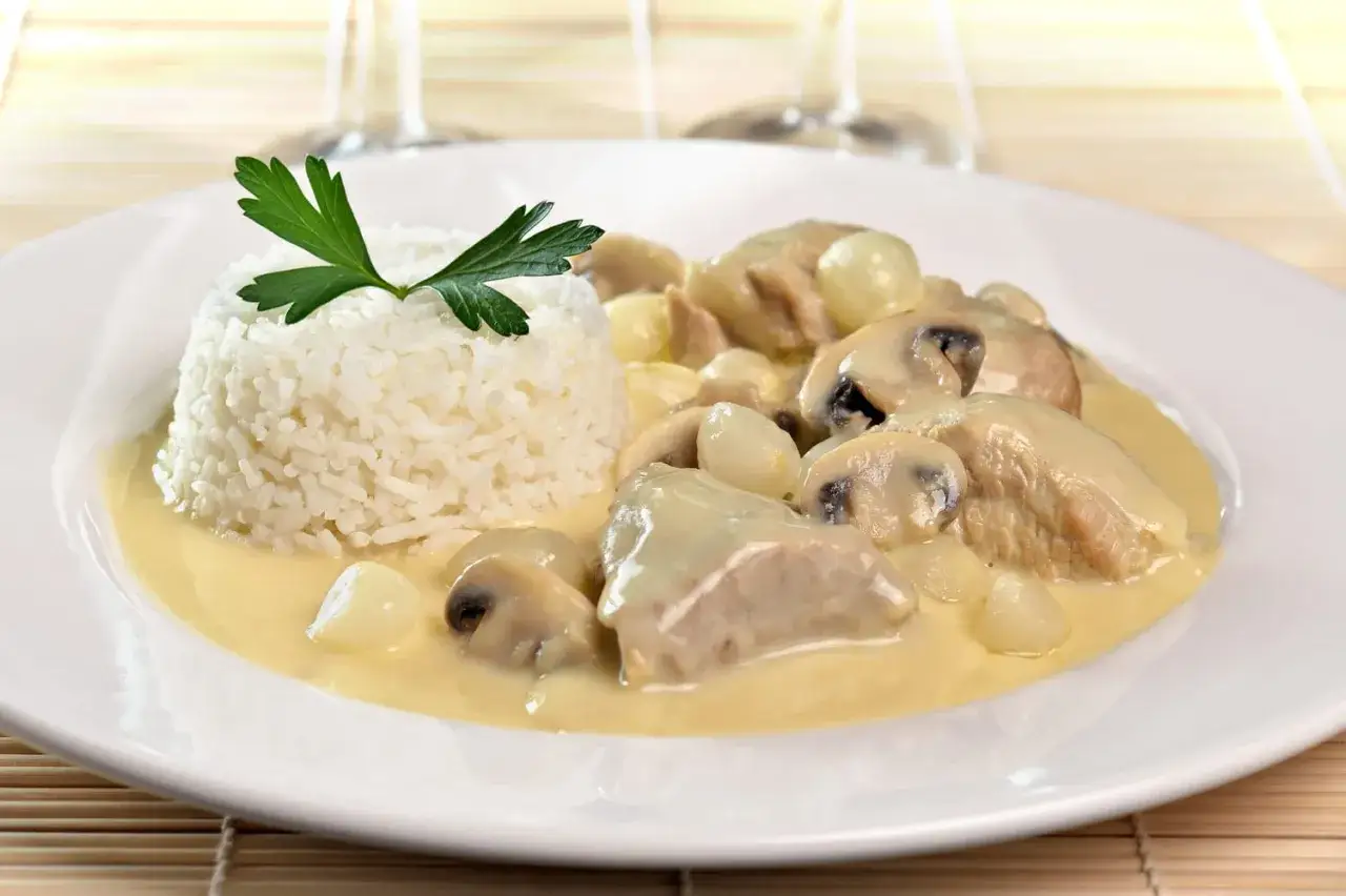Une assiette de blanquette de veau crémeuse avec des champignons et des oignons, servie avec une timbale de riz blanc garnie de persil.