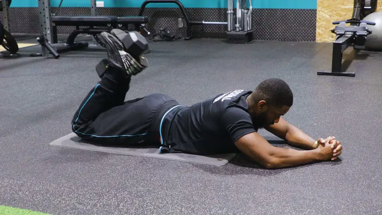 Mężczyzna wykonuje ćwiczenie hamstring curl z obciążeniem na kostkach, leżąc na macie w siłowni.